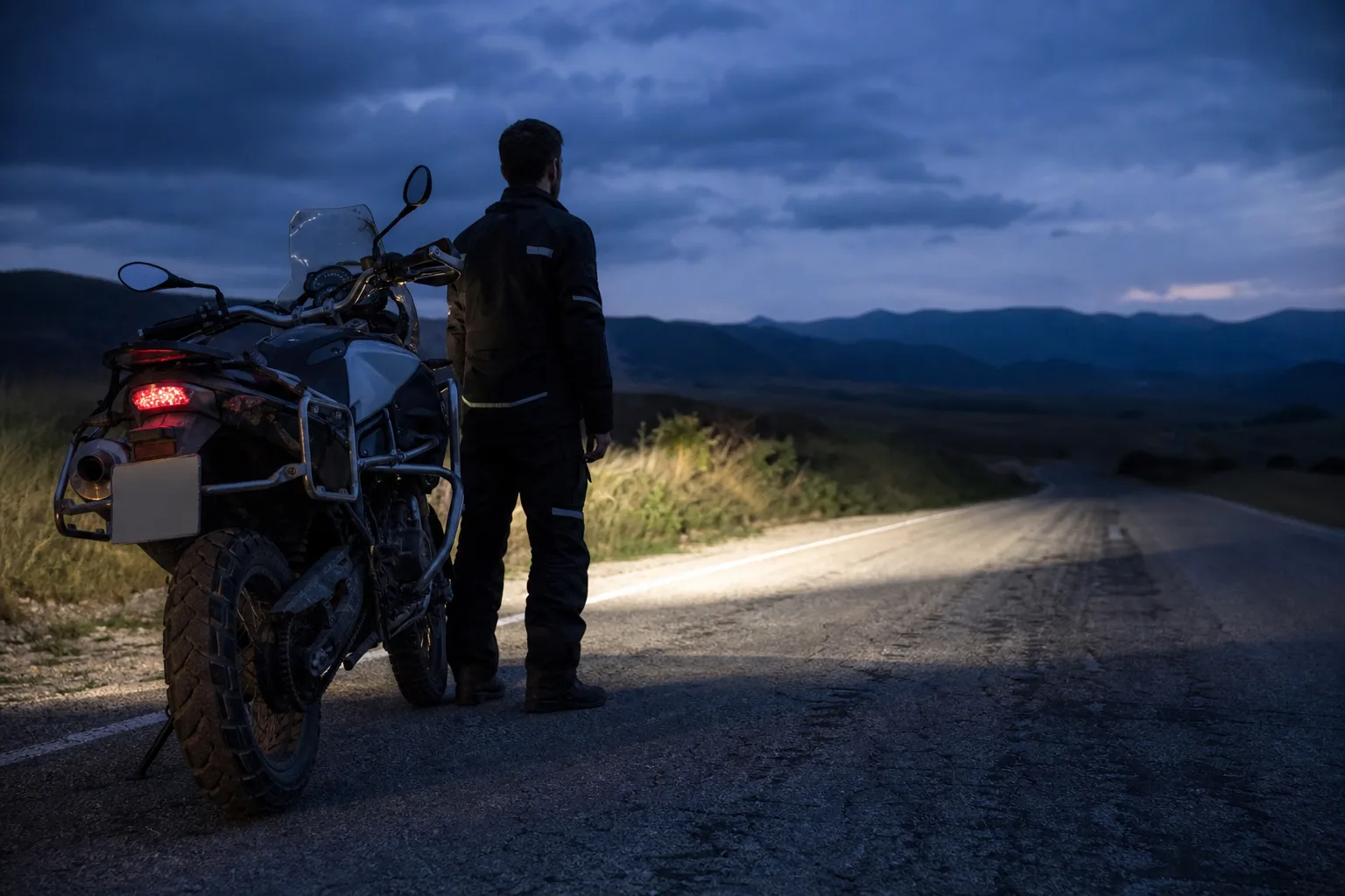 Motorcyclist assessing a motorcycle headlight at dusk on a quiet road.